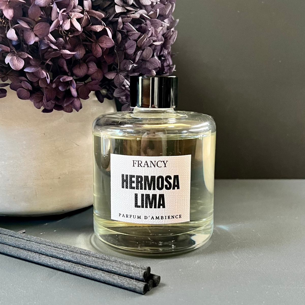 A clear glass reed diffuser with a label reading 'HERMOSA LIMA PARFUM D'AMBIENCE'. There are gray reeds at the bottom and a vase with purple flowers in the background.