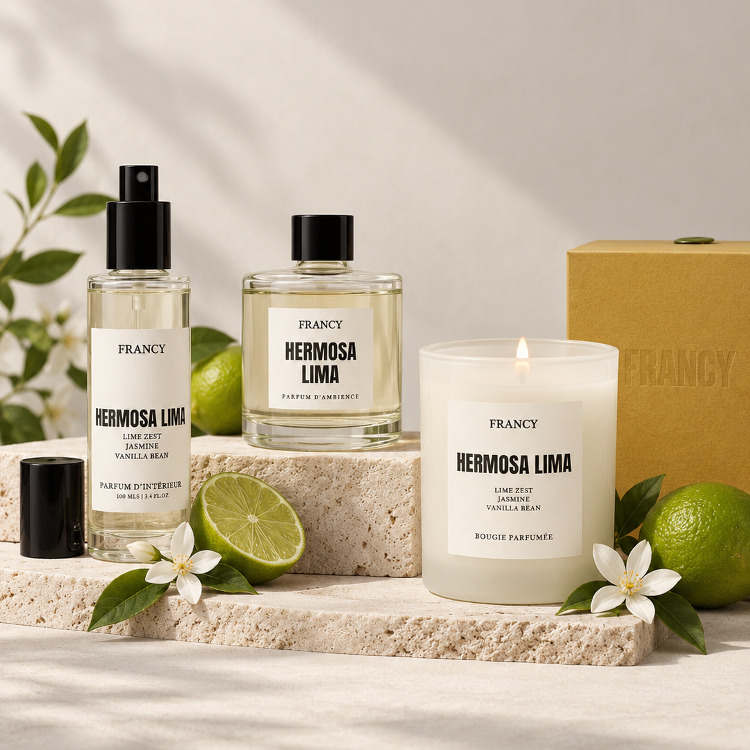 A fragrance set from the brand FRANCY, featuring a Hermosa Lima Eau de Parfum spray bottle, a matching perfume bottle, a lit candle, and a box, styled with limes, white flowers, and a stone slab against a sunlit neutral background.