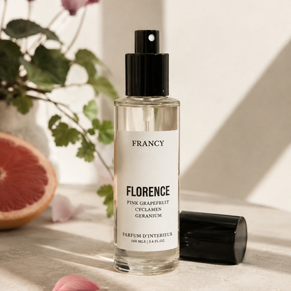 A clear glass perfume bottle with a black spray top and a white label reading FRANCY FLORENCE, placed on a stone surface beside its black cap, with a grapefruit slice, pink flower, and green leaves in the background.