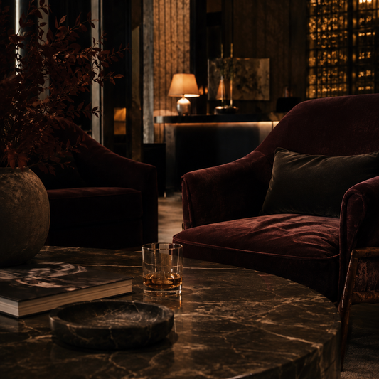 A moody, dimly lit lounge room featuring plush velvet armchairs in deep burgundy and a dark marble coffee table in the foreground, with warm ambient lighting and a blurred background.