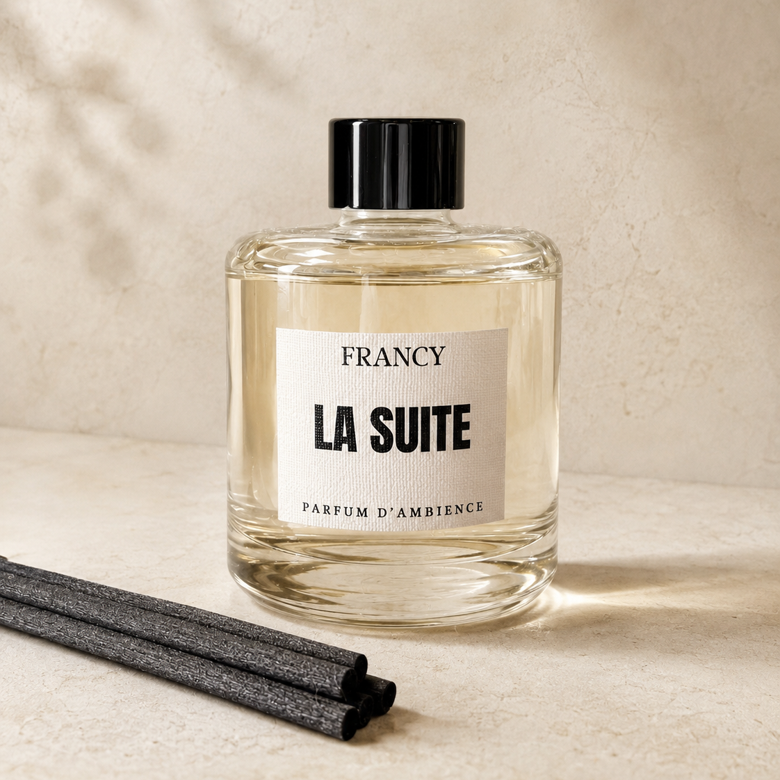 A clear glass bottle of Francy La Suite ambient fragrance with a black cap and white label, set on a beige surface with two dark reed sticks in the foreground and soft leaf shadows on the wall.