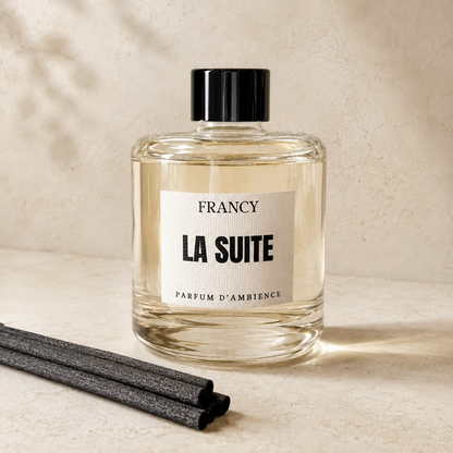 A clear glass bottle of Francy La Suite ambient fragrance with a black cap and white label, set on a beige surface with two dark reed sticks in the foreground and soft leaf shadows on the wall.