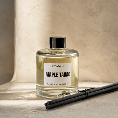 A clear glass reed diffuser bottle labeled 'FRANCY MAPLE TABAC' with a black cap, set on a stone surface against a textured beige wall. Two dark reeds rest beside it in soft sunlight.