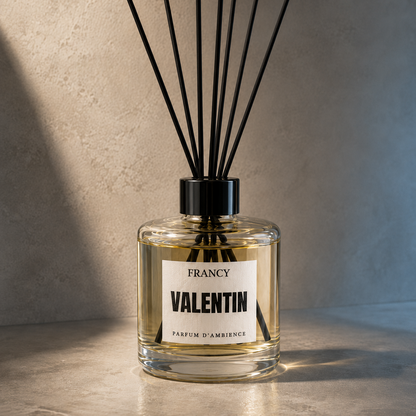A clear glass reed diffuser bottle with a black cap and black reeds, labeled 'FRANCY VALENTIN PARFUM D'AMBIANCE', set on a stone-like surface against a textured wall with dramatic warm lighting.